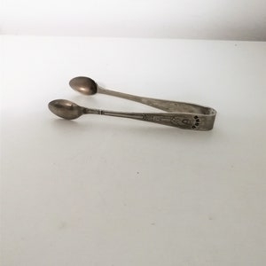 May include: Silver-toned sugar tongs with spoon-shaped ends. The tongs have a decorative design on the handle. The image is set against a white background.