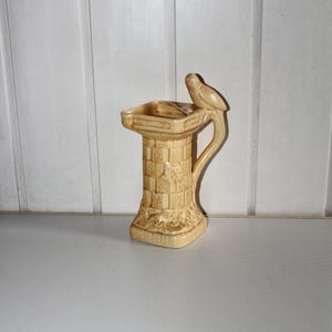 May include: A beige ceramic bird bath with a bird perched on the side. The bird bath has a square base and a cylindrical body with a textured brick pattern. The top has a rectangular basin.