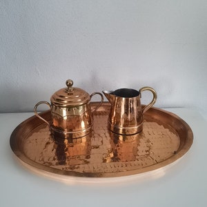 May include: A copper tea set with a hammered texture, including a lidded sugar bowl and a creamer, sits on a matching oval tray. The set has a warm, metallic sheen, perfect for serving tea or coffee.
