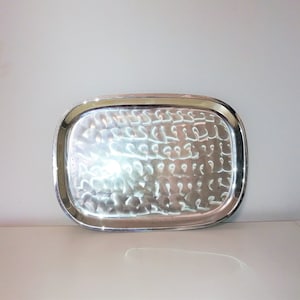 May include: A rectangular silver serving tray with rounded corners. The tray has a hammered texture on the inside and a polished, reflective surface. The rim of the tray is smooth and shiny.