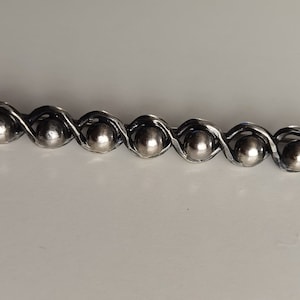 May include: A silver-toned barrette with a series of small, round, metallic beads. The beads are connected by a twisted metal design, creating a decorative and elegant accessory. The barrette is set against a white background.