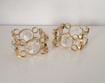 May include: Two gold-colored candle holders with clear, faceted crystal accents. Each holder features a circular design with multiple small rings and a large, central crystal. The holders are placed on a white surface.