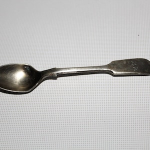 Peut inclure: Une petite cuillère en argent antique avec un bol arrondi et un manche rectangulaire. La cuillère est ternie, montrant des signes d'âge et d'utilisation. La poignée porte une petite inscription. La cuillère est probablement pour servir ou manger.