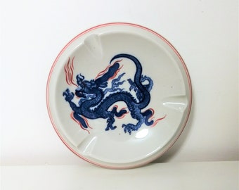 May include: White ceramic ashtray with a blue dragon design. The dragon is detailed with red accents and sits on a white background. The ashtray has a red rim and three cigarette rests.