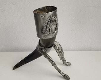 May include: A silver-colored, horn-shaped drinking vessel with a black horn base and three textured legs. The vessel has an embossed design around the top. The object is on a white surface.