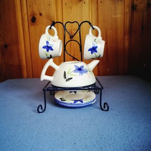 May include: A white ceramic tea set with blue floral designs, including a teapot, two cups, and a saucer, displayed on a black metal stand. The set is placed on a blue surface against a wooden background.