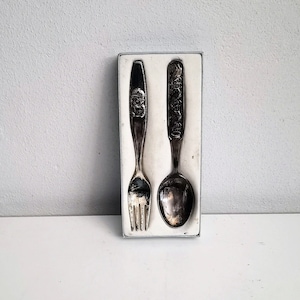 Pode incluir: Um conjunto de talheres de bebé de prata numa caixa branca. O garfo e a colher têm um design decorativo.