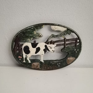 Puede incluir: Pieza decorativa ovalada de hierro fundido con una vaca en blanco y negro en un paisaje de pasto. La vaca está cerca de una valla de madera y follaje verde. El marco es verde oscuro con detalles marrones y blancos.