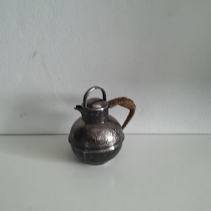 Puede incluir: Una pequeña tetera plateada con cuerpo redondeado y asa trenzada. La tetera tiene tapa y boquilla. La tetera está sobre una superficie blanca.