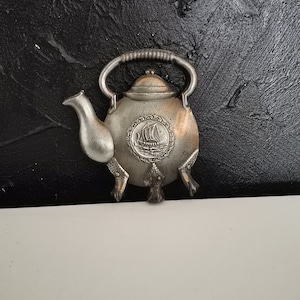 May include: A silver-colored metal teapot-shaped ornament with a handle, spout, and three feet. The teapot has a circular emblem with a ship design. The background is a textured black surface.