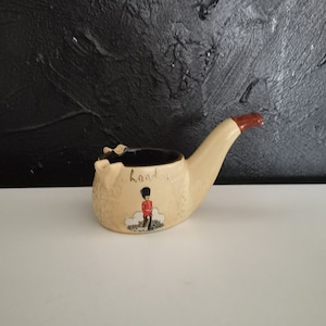 May include: A beige ceramic pipe-shaped ashtray with a black interior. The ashtray has a red tip and a painted illustration of a London guard. The word "London" is written on the side.