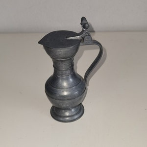Könnte beinhalten: Kleine, antike Zinnkanne mit Klappdeckel und gebogenem Griff. Die Kanne hat einen bauchigen Körper mit horizontalen Rippen und einem kleinen Ausguss. Der Deckel hat einen kleinen Knopf zum Öffnen. Zum Servieren von Getränken.