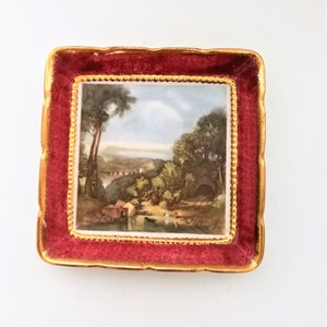 May include: Square decorative dish with a landscape painting. The dish has a red border with a gold trim and a gold edge. The painting depicts a river scene with trees, a bridge, and buildings.