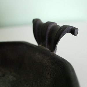 May include: Close-up of a dark, possibly antique, metal object. The object has a textured surface and a curved handle with a rounded end. The handle has a decorative, ridged design.