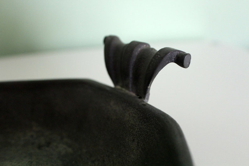 May include: Close-up of a dark, possibly antique, metal object. The object has a textured surface and a curved handle with a rounded end. The handle has a decorative, ridged design.