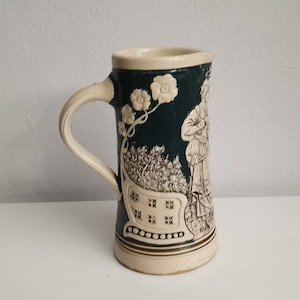 May include: A ceramic beer stein with a handle, featuring a dark green background and cream-colored relief carvings. The carvings depict figures and floral designs. The stein is approximately 6 inches tall.