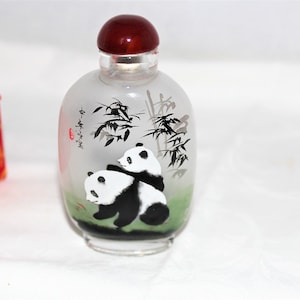 Könnte beinhalten: Eine klare Glasflasche mit einem dunkelroten Verschluss. Die Flasche hat ein handgemaltes Design von zwei Pandas, Bambus und chinesischen Schriftzeichen. Die Flasche ist teilweise mit einem grünen und weißen Farbverlauf gefrostet.