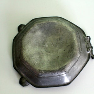 May include: A vintage, octagonal pewter dish with a dark, aged patina. The dish has decorative handles and feet, suggesting a historical or antique aesthetic. The surface shows signs of wear, adding to its character.