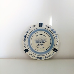 Puede incluir: Un cenicero redondo de cerámica con borde festoneado. El cenicero presenta un diseño floral azul y blanco, una imagen central y texto. Tiene cuatro reposos integrados para cigarrillos.