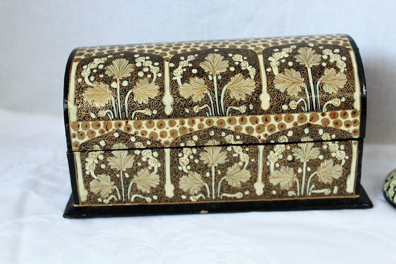 May include: A decorative, arched-top box with a black base and lid. The box is adorned with a repeating pattern of stylized leaves in cream and gold against a dark brown background. The design includes small, circular accents.