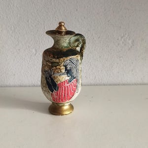 Può includere: Un piccolo vaso decorativo in ceramica con coperchio e manico. Il vaso presenta una base e un coperchio dorati, con un disegno dipinto di una figura in rosso e nero. Il vaso è alto circa 15 cm.