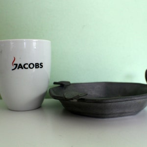 May include: A white ceramic mug with the word "JACOBS" printed in black and red, next to a dark gray ashtray with a unique handle design. The ashtray is made of metal.