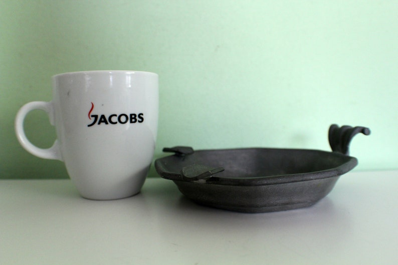 May include: A white ceramic mug with the word "JACOBS" printed in black and red, next to a dark gray ashtray with a unique handle design. The ashtray is made of metal.