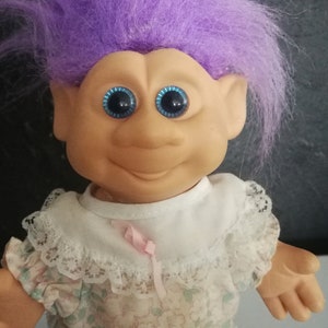May include: A vintage Troll doll with vibrant purple hair and large blue eyes. The doll has a smiling expression and is wearing a floral print dress with a white lace collar and a pink ribbon.