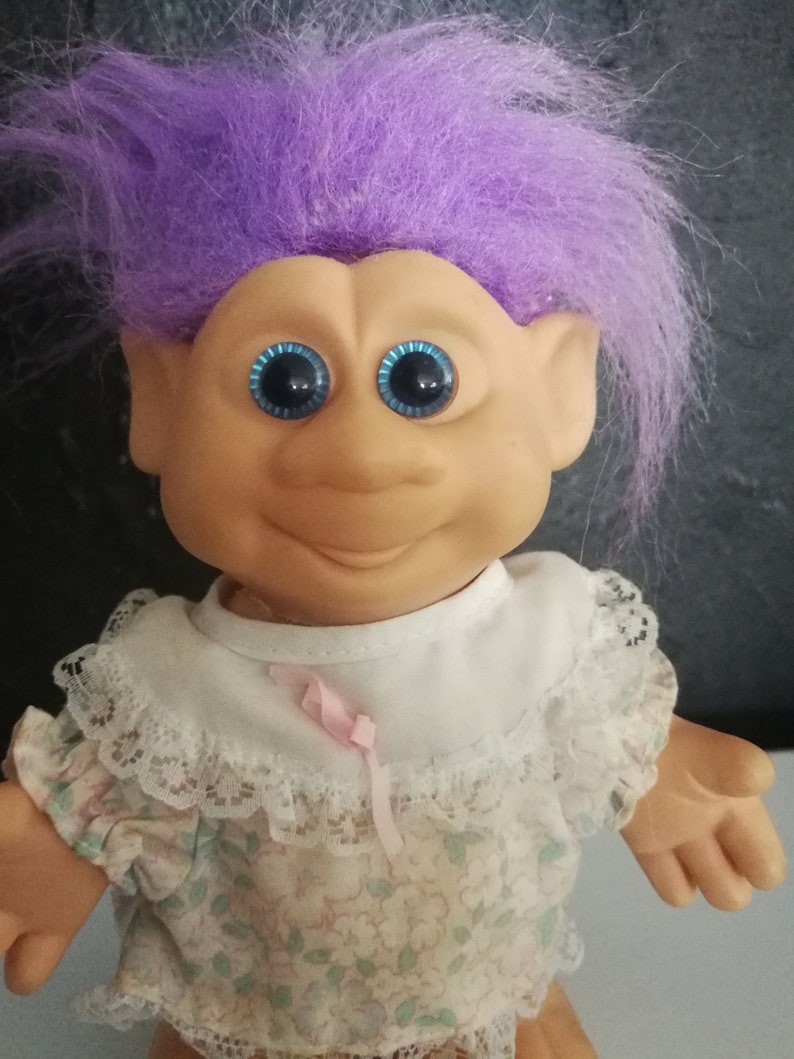 May include: A vintage Troll doll with vibrant purple hair and large blue eyes. The doll has a smiling expression and is wearing a floral print dress with a white lace collar and a pink ribbon.