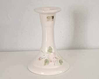 May include: Cream-colored ceramic candlestick holder with a flared base and a wide top. The base is decorated with pink floral and green leaf accents. A small sticker is visible on the top.