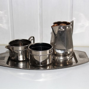 May include: A silver-toned metal tea service set consisting of a small creamer, a sugar bowl, and a tall pitcher, all arranged on a matching rectangular tray with rounded corners and decorative cut-out handles on each side.