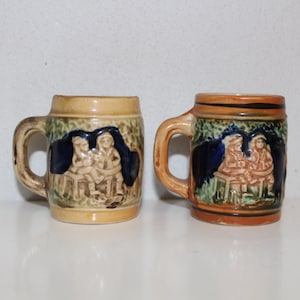 Puede incluir: Dos pequeñas tazas de cerámica con diseños pintados en marrón, verde y azul. Cada taza presenta una escena de tres personas sentadas en una mesa.