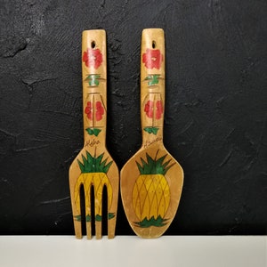 Könnte beinhalten: Holz-Gabel und Löffel-Set mit tropischem Thema. Gabel und Löffel sind mit einem gelben Ananas-Design und roten Blumenakzenten bemalt. Das Wort "Aloha" steht auf der Gabel und "Hawaii" auf dem Löffel.