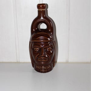 May include: A brown ceramic bottle with a face design. The bottle has a handle and a stopper. The face has detailed features. The text "PAZ-SOLDA PISCO" is at the base. A decorative item.