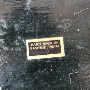 May include: Close-up of a dark, textured surface with scratches and wear. A rectangular sticker with the text "HAND MADE IN KASHMIR (INDIA)" is affixed to the surface. The surface appears to be painted or stained.