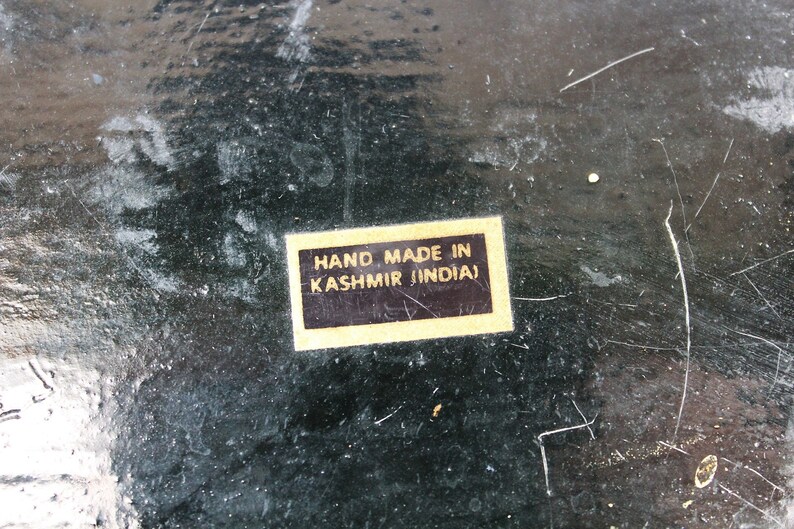 May include: Close-up of a dark, textured surface with scratches and wear. A rectangular sticker with the text "HAND MADE IN KASHMIR (INDIA)" is affixed to the surface. The surface appears to be painted or stained.