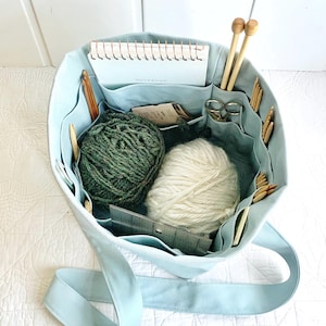 May include: A pale blue tote bag containing knitting and crochet supplies. It features two balls of yarn, one green and one white, plus knitting needles, crochet hooks, scissors, a notebook, and a metal ruler. The bag has several pockets for storage.