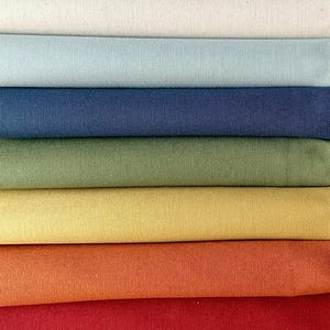 May include: A selection of fabric rolls in a range of colours. The colours displayed are cream, pale blue, navy, olive green, yellow, orange, and red. The fabric has a textured finish, possibly canvas or a similar material.
