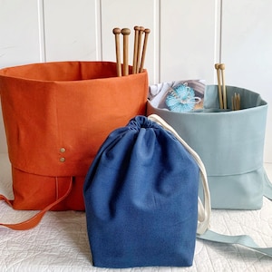 May include: Three fabric storage containers in shades of orange, blue, and light blue. The orange container holds wooden knitting needles. The blue drawstring bag is in front. The light blue container holds knitting supplies. The containers have straps.