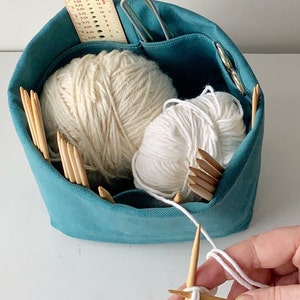 Small blue knitting bag with cream and white yarn.  Hands in lower right corner are knitting from this yarn.