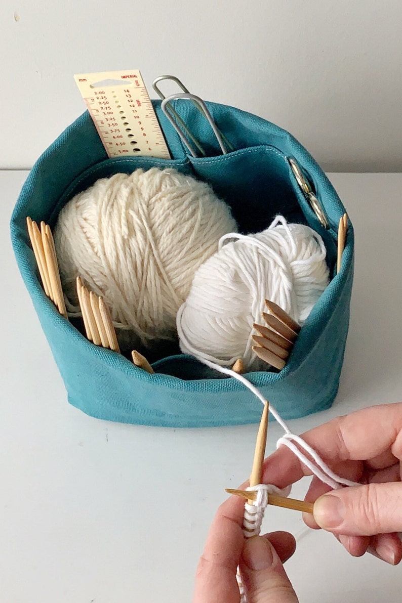 Small blue knitting bag with cream and white yarn.  Hands in lower right corner are knitting from this yarn.