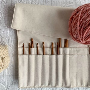 May include: A cream canvas crochet hook holder with wooden hooks. Two balls of yarn, one cream and one pink, are next to the holder. The holder is rolled up and has pockets to hold the hooks. The yarn is measured in grams.