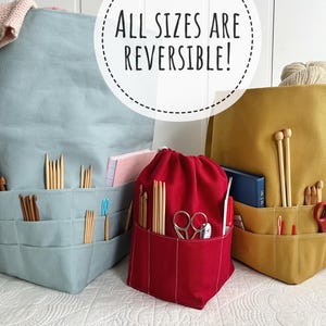 May include: Three fabric project bags in blue, red, and mustard yellow, each with multiple pockets. The blue bag has a folded knitted item on top. The red bag is a drawstring bag. The text "ALL SIZES ARE REVERSIBLE!" is in the image.