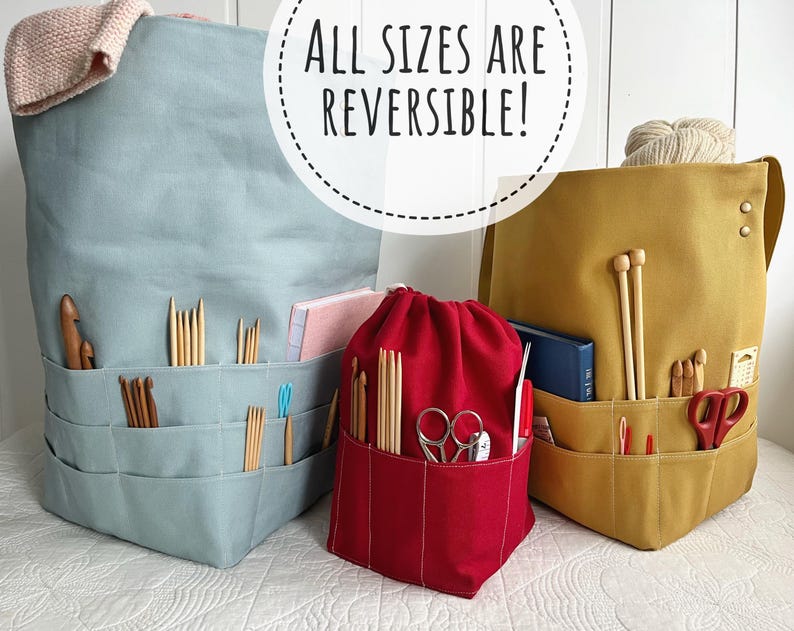 May include: Three fabric project bags in blue, red, and mustard yellow, each with multiple pockets. The blue bag has a folded knitted item on top. The red bag is a drawstring bag. The text "ALL SIZES ARE REVERSIBLE!" is in the image.