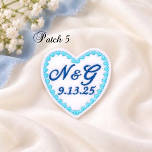May include: A white heart-shaped embroidered patch featuring a bright blue scalloped border. The center displays the dark blue embroidered letters N & G above the date 9.13.25. The text Patch 5 is visible in the upper left corner.