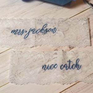 Op de afbeelding: Twee witte kanten kousenbanden met blauw geborduurde tekst. Op &eacute;&eacute;n kousenband staat "mrs. jackson" en op de andere "nice catch". Een zilveren schaar staat op de achtergrond, samen met een klosje garen.