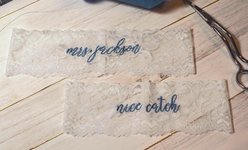 Op de afbeelding: Twee witte kanten kousenbanden met blauw geborduurde tekst. Op &eacute;&eacute;n kousenband staat "mrs. jackson" en op de andere "nice catch". Een zilveren schaar staat op de achtergrond, samen met een klosje garen.