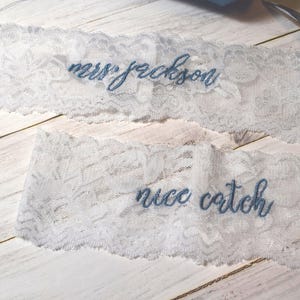 Op de afbeelding: Twee witte kanten kousenbanden met blauw geborduurde tekst. Op &eacute;&eacute;n kousenband staat "mrs jackson" en op de andere "nice catch". De kousenbanden liggen op een licht houten oppervlak.