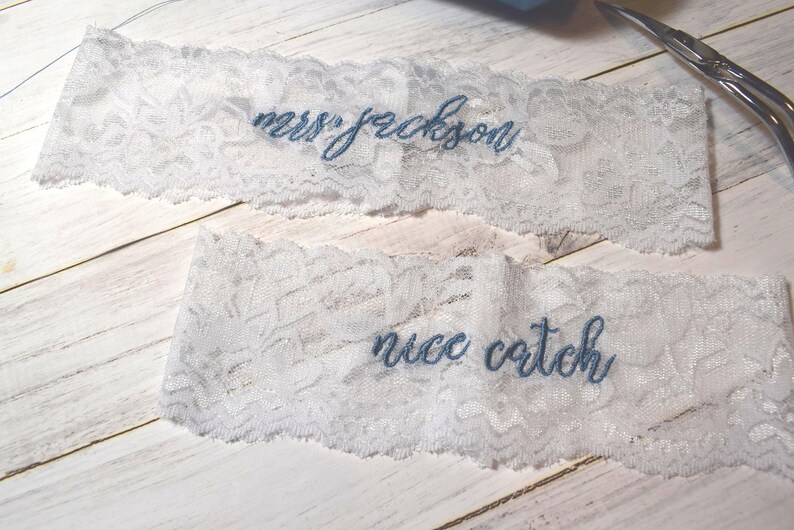 Op de afbeelding: Twee witte kanten kousenbanden met blauw geborduurde tekst. Op &eacute;&eacute;n kousenband staat "mrs jackson" en op de andere "nice catch". De kousenbanden liggen op een licht houten oppervlak.