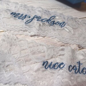 Op de afbeelding: Twee stukken wit kant met blauw geborduurde tekst. Op &eacute;&eacute;n stuk staat "mrs. jackson" en op het andere "nice catch". Het kant ligt op een licht houten oppervlak.